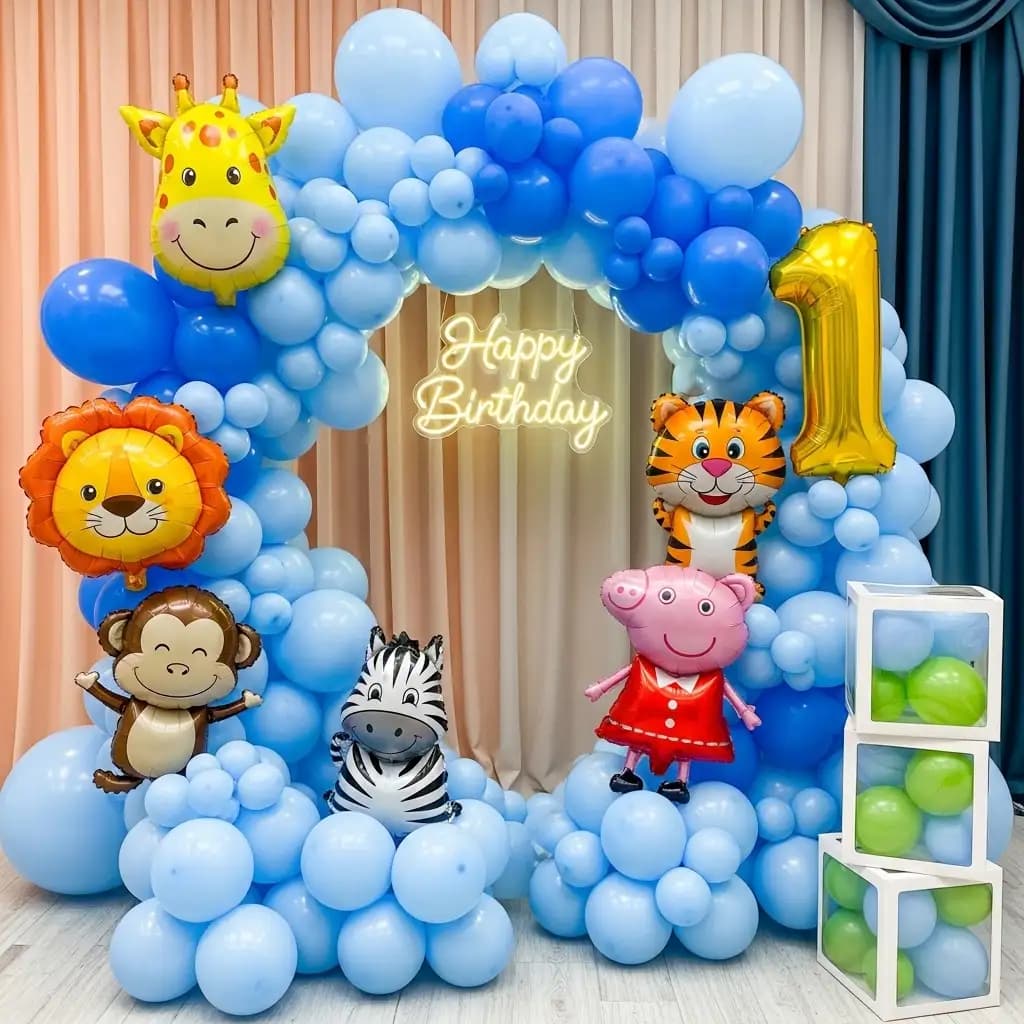 Jungle-themed first birthday setup featuring a vibrant blue balloon arch adorned with animal-shaped balloons like a giraffe, lion, monkey, and zebra. A glowing “Happy Birthday” sign and golden “1” balloon highlight the celebration, with green balloon-filled boxes adding a playful touch.