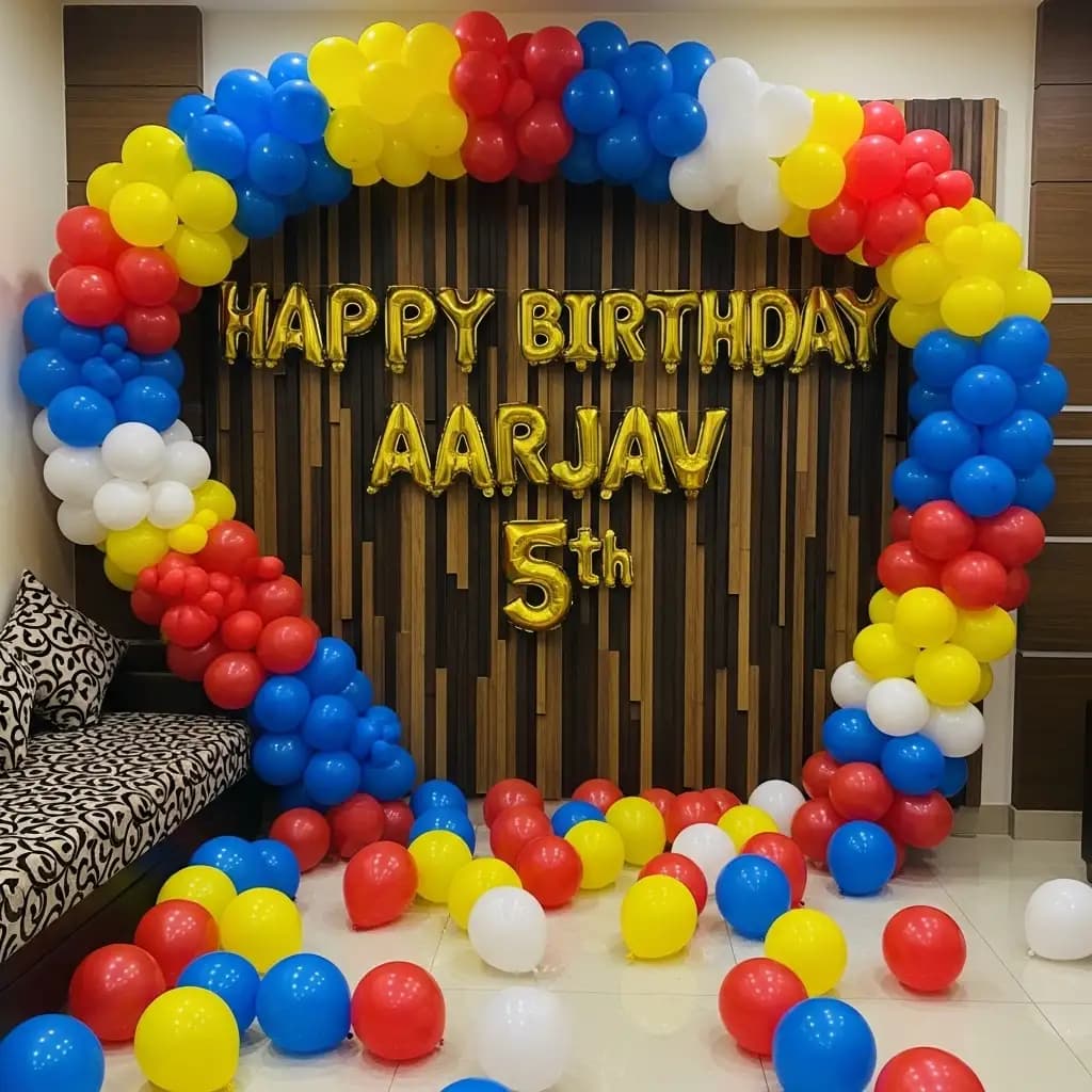 Vibrant 5th birthday setup featuring a multicolor balloon arch in blue, red, yellow, and white, gold balloon letters spelling “HAPPY BIRTHDAY AARJAV 5th,” and a cozy seating area with patterned cushions for a fun, festive celebration.